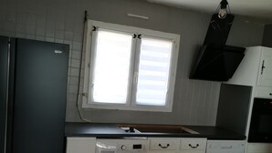 Eine kleine Küche mit Waschmaschine, Kühlschrank, einem Fenster mit Jalousien und einer Pendelleuchte.