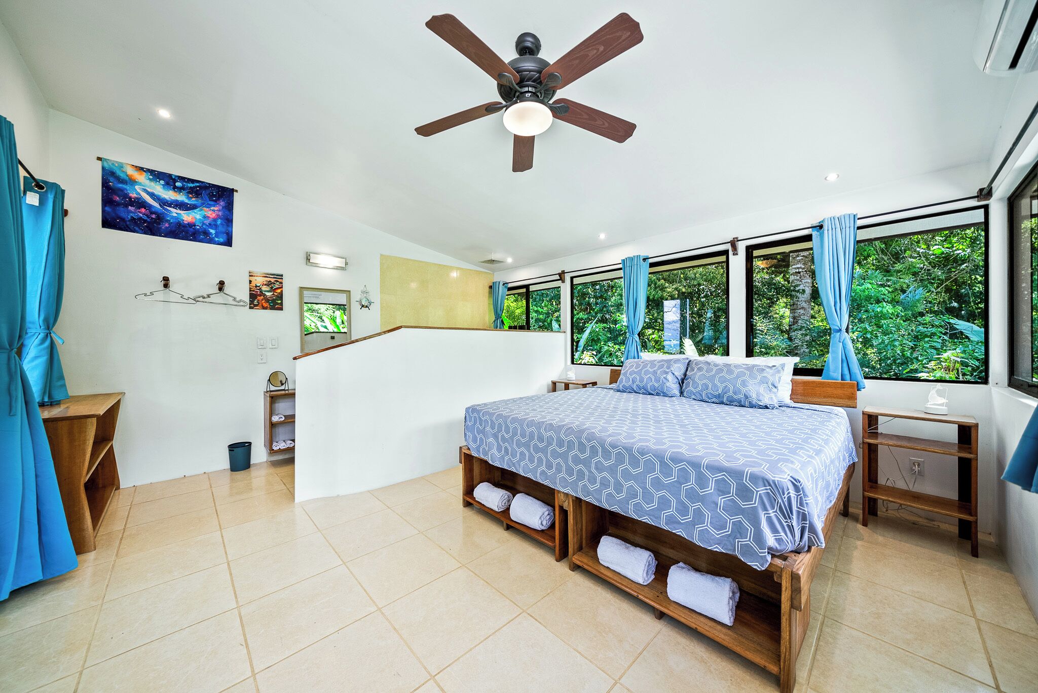 Une chambre à coucher comprenant un lit, un ventilateur de plafond, une fenêtre avec des rideaux, un escalier et une œuvre d’art accrochée au mur.