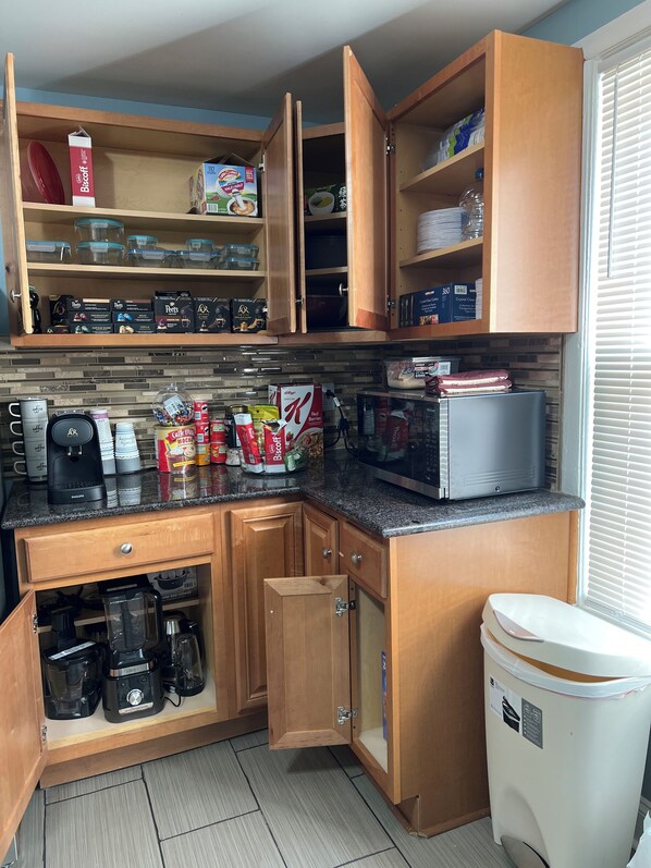 A compact kitchen with wooden cabinets, a microwave, coffee maker, and various food items.