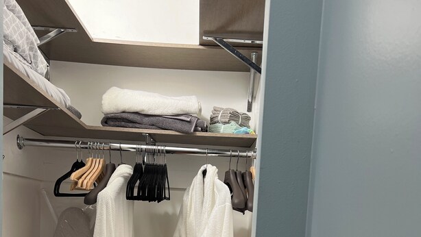 Un dressing où l’on peut entrer, avec une tringle à vêtements, une étagère où sont pliées des serviettes et un peignoir blanc accroché à la porte.