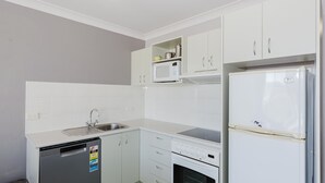 A compact kitchen with white cabinetry, a built-in microwave, a stainless steel sink, a dishwasher, and a white refrigerator. - Horizons - 418/10 Kosciuszko Road (Jindabyne)