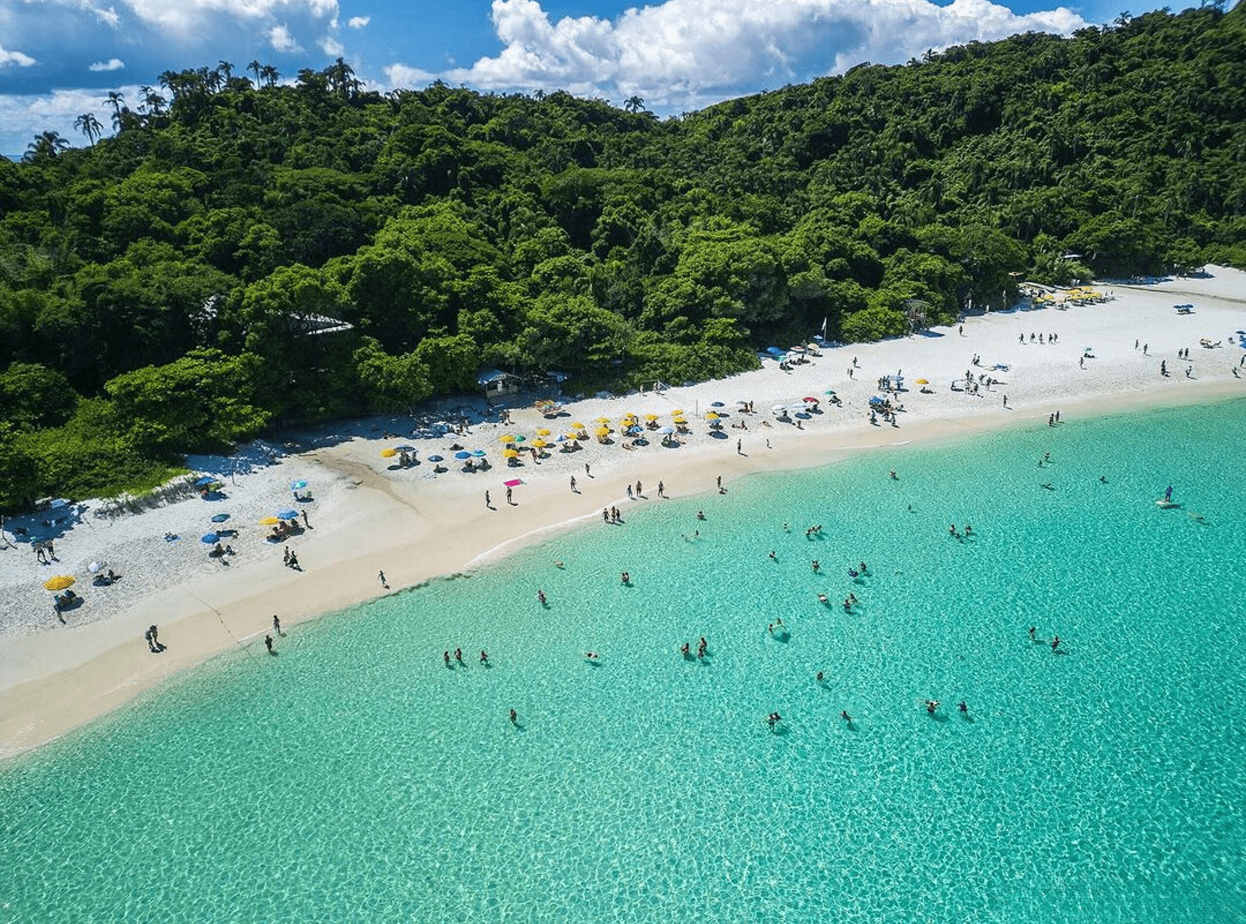 Luchtbeeld van een strand met helder turquoise water, een zandstrand en groene heuvels op de achtergrond.