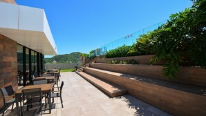 An outdoor patio with wooden tables and chairs, a glass railing, and a view of the hills. - Studio Shopping OKA Floripa - Campeche - SOK125 (Florianópolis)