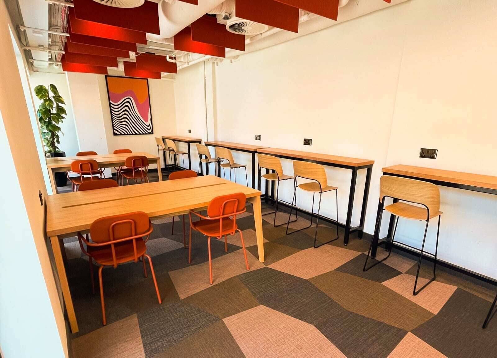 Une salle de réunion moderne, dotée de longues tables et de chaises, d’une grande plante et d’un tableau abstrait aux couleurs vives accroché au mur.