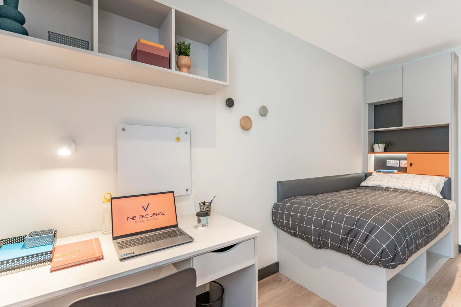 Ein modernes Schlafzimmer mit einem Bett, einem Schreibtisch mit Laptop, einer Whiteboard-Tafel und Regalen mit Büchern und einer Pflanze.
