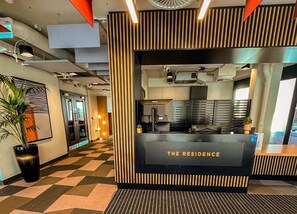 Un comptoir d’accueil avec une enseigne indiquant « LA RÉSIDENCE », situé dans un hall d’entrée moderne doté d’un sol à motifs géométriques et agrémenté d’une plante en pot.