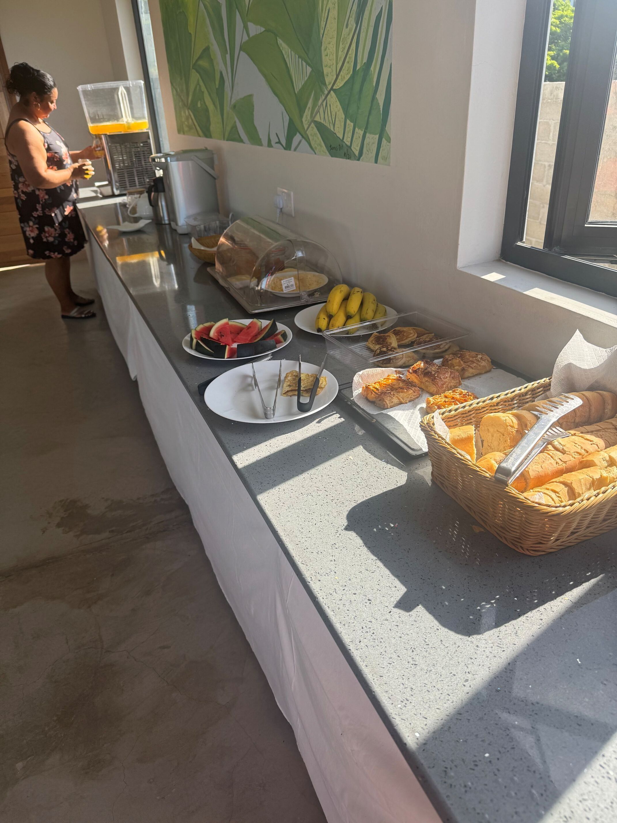 Una mesa de buffet con diversas opciones de comida, como frutas, pasteles y pan, dispuesta sobre un mostrador con una ventana al fondo.