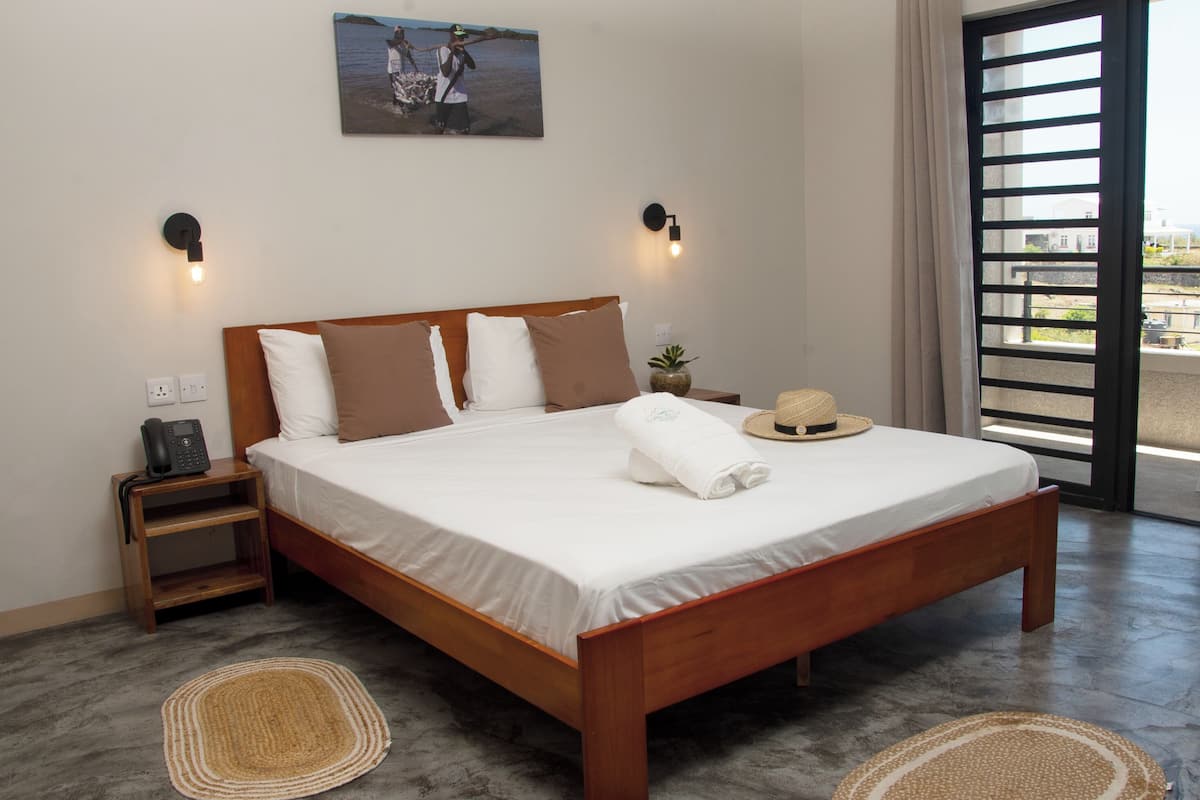 A bedroom with a wooden bed, white bedding, two wall-mounted lights, a small nightstand, a telephone, a window with a view, and a picture on the wall.