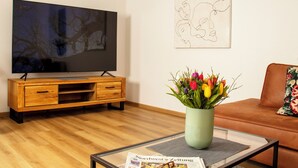 A living room with a wooden TV stand, a flat-screen TV, a vase with flowers, a newspaper, and a couch with a floral pillow.