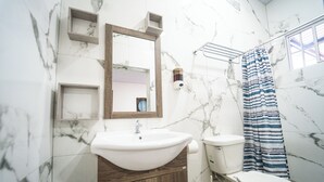 Une salle de bain avec un mur en marbre, un meuble-lavabo en bois, un lavabo blanc, des toilettes et un rideau de douche.