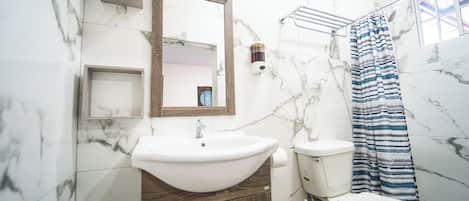 A bathroom with a marble wall, a wooden vanity, a white sink, a toilet, and a shower curtain.