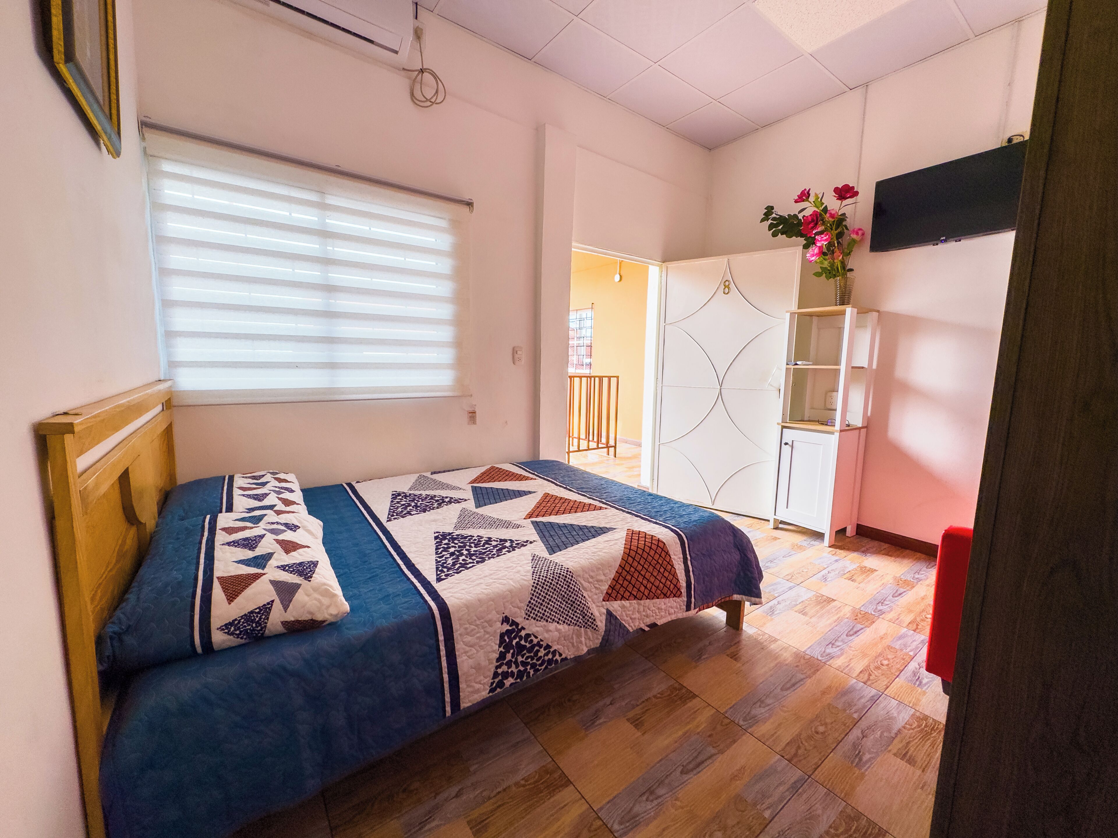 Une chambre à coucher avec un lit, une fenêtre avec des stores, une armoire et un téléviseur fixé au mur.