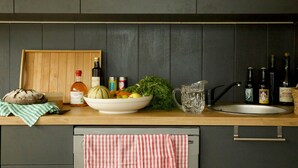 Un plan de travail de cuisine avec une miche de pain, un saladier de légumes, une bouteille d’huile d’olive et plusieurs bouteilles de vin.