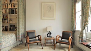 Une pièce comprenant une bibliothèque, deux chaises en bois, une petite table avec une lampe et un tableau encadré accroché au mur.