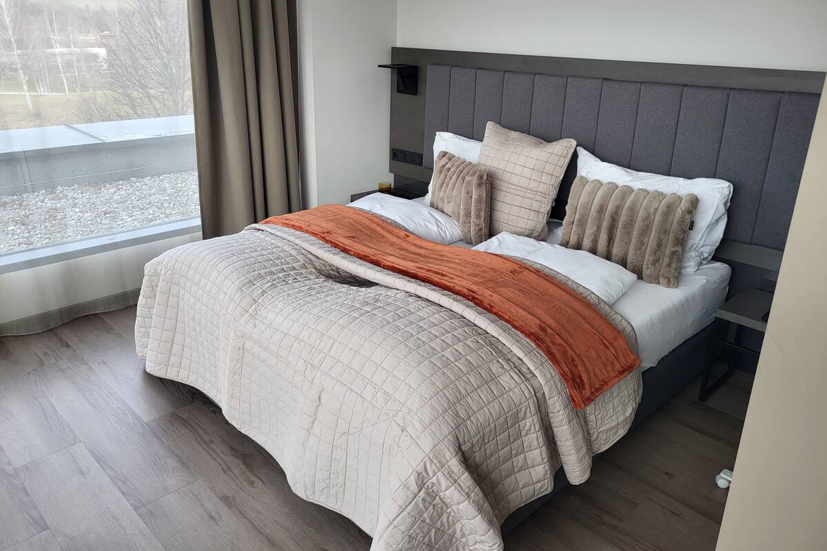 A neatly made bed with a quilted cover, a grey headboard, and a view of a lake through the window.