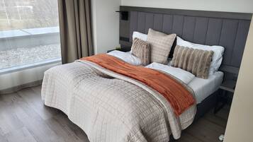 A neatly made bed with a quilted cover, a grey headboard, and a view of a lake through the window.