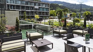 An outdoor patio with seating, a water feature, and a view of a modern building and mountains. - DION Hotel Wattens (Wattens)