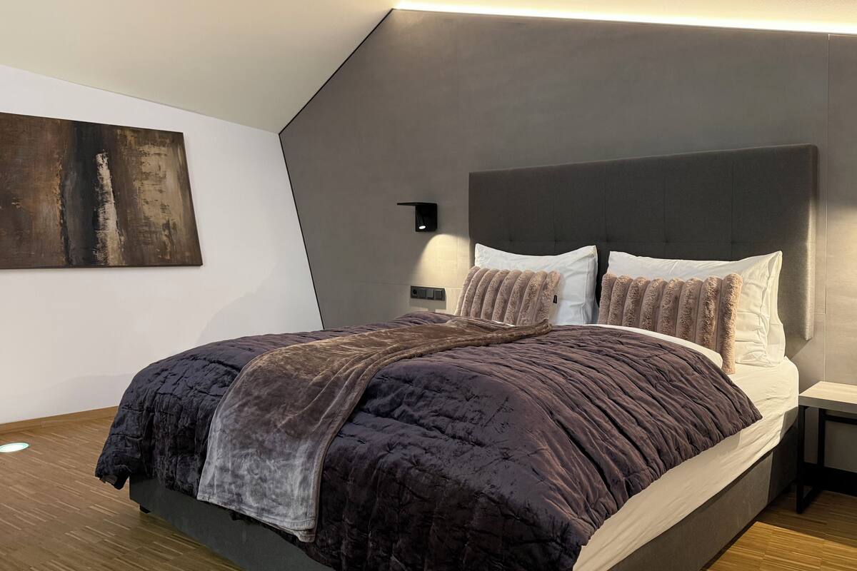A modern bedroom with a large bed, a headboard, bedside tables, and a wall-mounted light fixture.