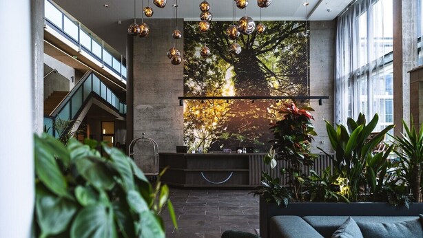 A lobby with a reception desk, a staircase, and a large window with a view of trees. - DION Hotel Wattens (Wattens)