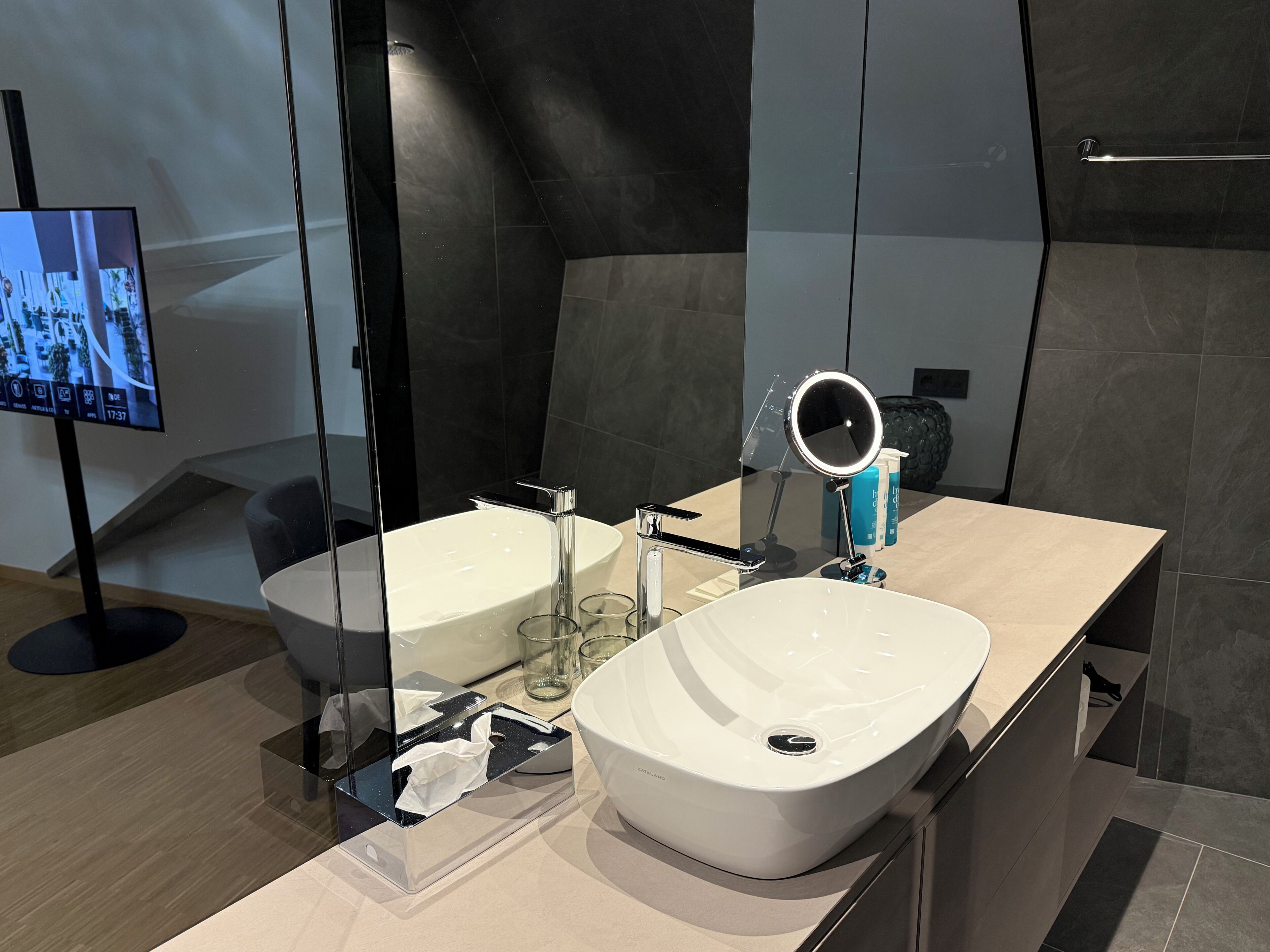 A modern bathroom with a large mirror, two sinks, a vanity, and a television.