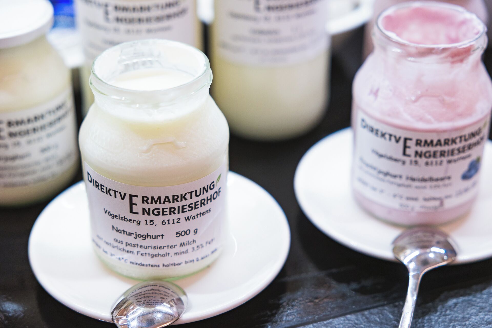 Jars of dairy products on plates with labels, a spoon, and a dark surface.