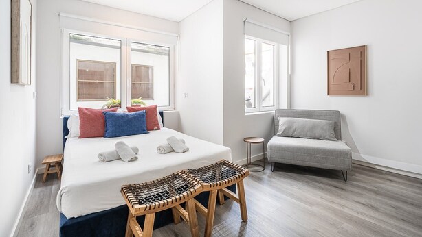 Ein modernes Schlafzimmer mit einem großen Bett, einem grauen Sofa, einem kleinen Tisch und einem Fenster mit Blick.