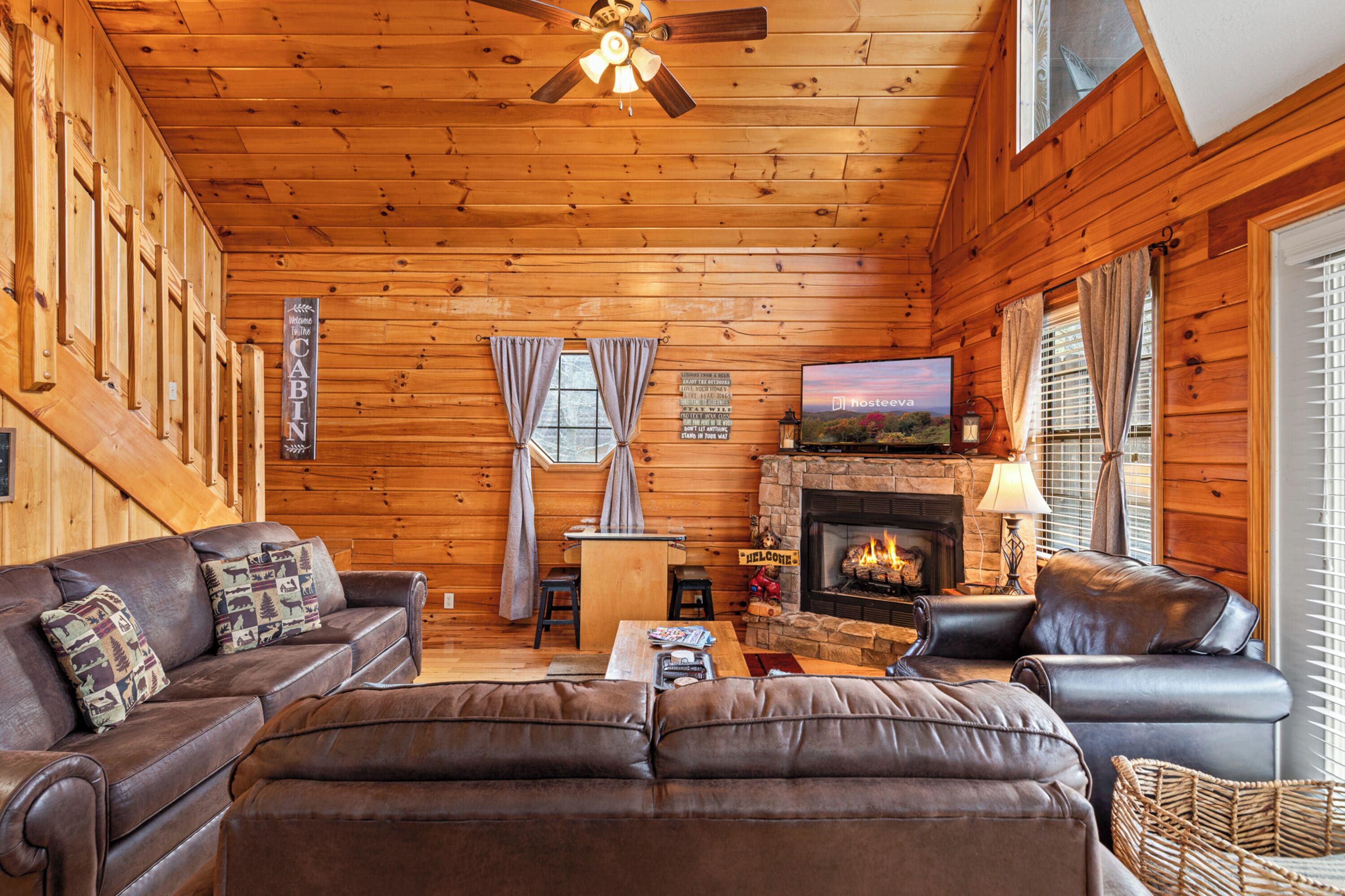 A log cabin interior with a fireplace, leather sofas, a television, and a ceiling fan.