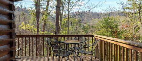 A wooden deck with a table and chairs, overlooking a forested area with buildings in the distance.