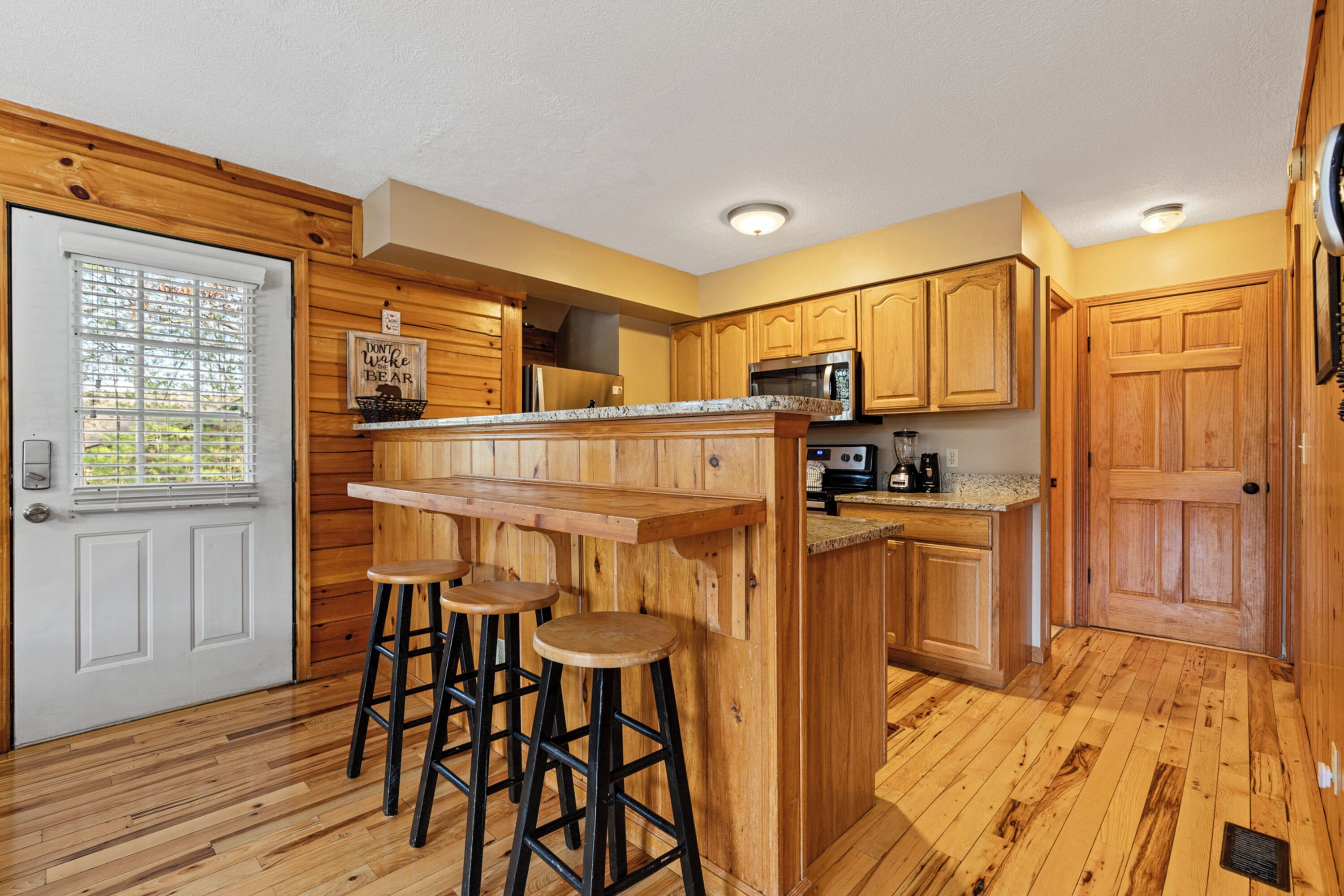 A kitchen with wooden cabinets, a countertop, a microwave, and a sink. There are three stools at the bar counter.