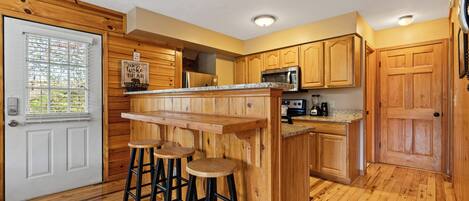 A kitchen with wooden cabinets, a countertop, a microwave, and a sink. There are three stools at the bar counter.