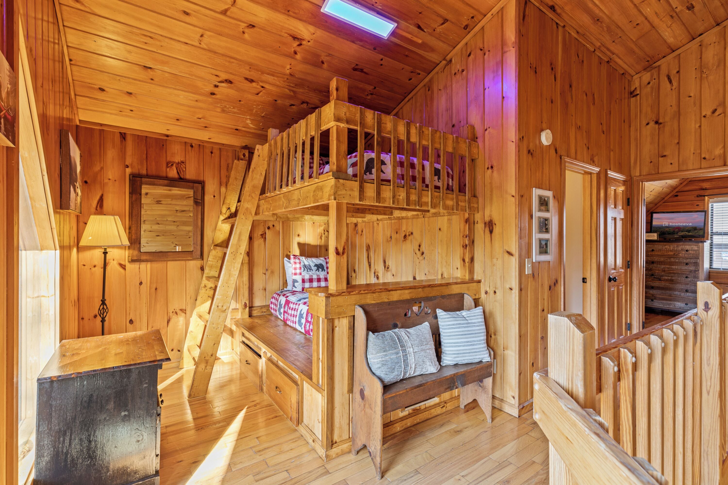 Interior de una cabaña de madera con una cama litera, un banco de madera con almohadones, un escritorio y una escalera.