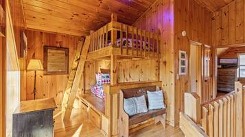 A wooden cabin interior with a bunk bed, a wooden bench with pillows, a desk, and a ladder.