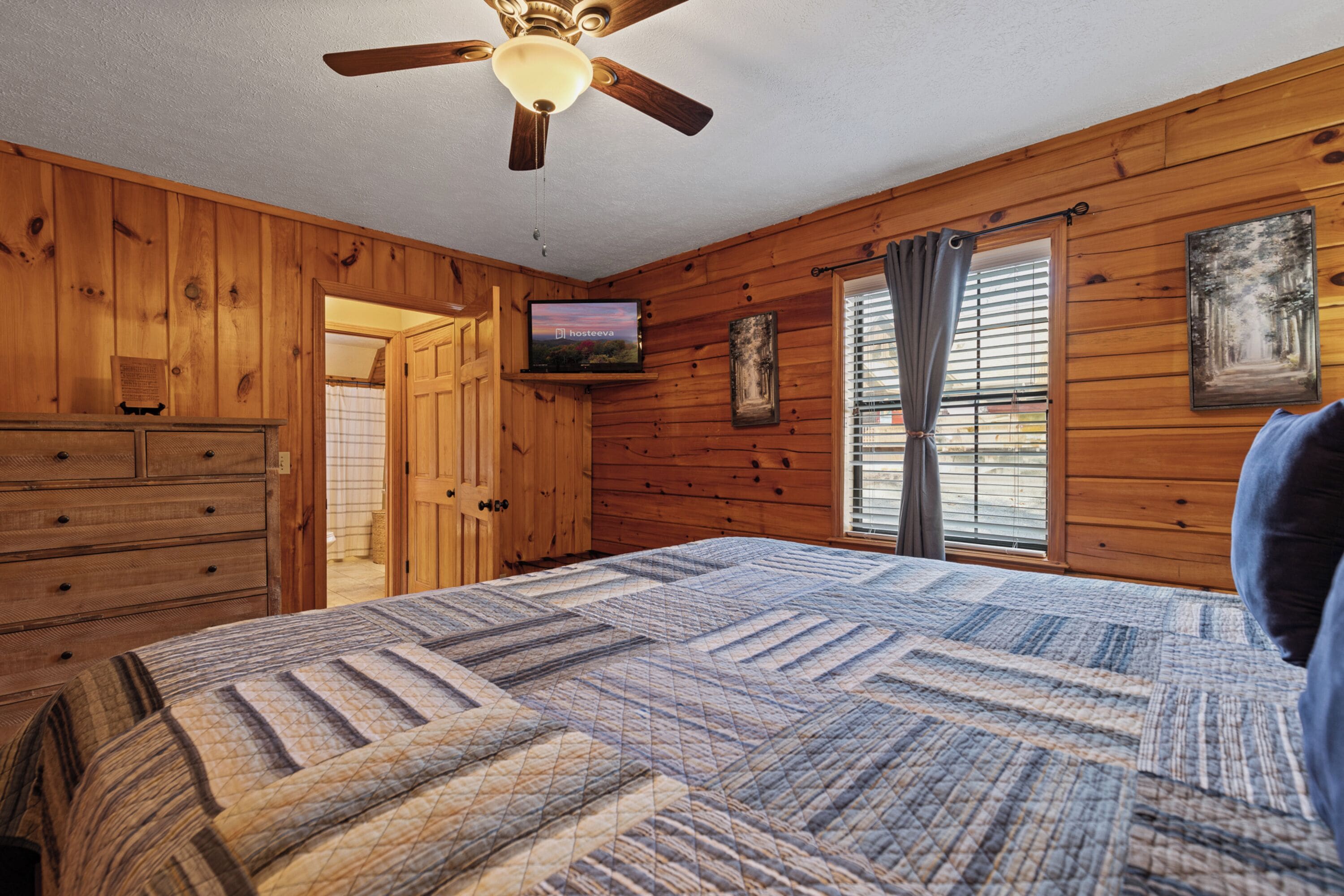 A wooden cabin bedroom with a bed, a ceiling fan, a television, and a window with curtains.