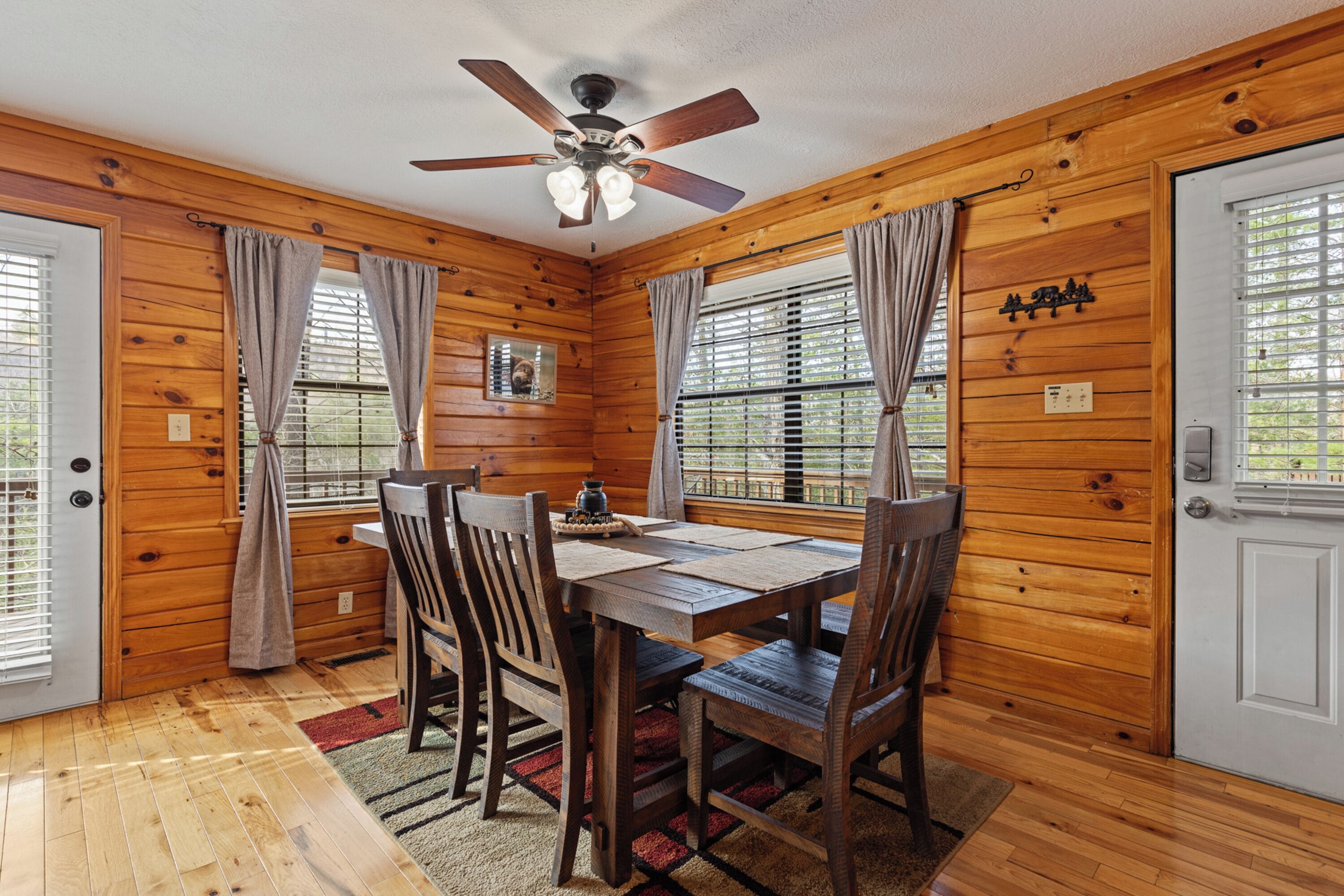 A wooden cabin interior with a dining table, chairs, a ceiling fan, and a window with curtains.