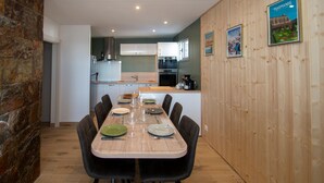 Un comedor con una larga mesa de madera dispuesta para cuatro personas, rodeada de sillas negras. Se ve una cocina con armarios blancos y una pared de piedra al fondo.