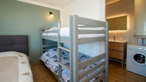 Una cama litera con cabecera, una lavadora, una mesita de noche pequeña y un espejo.