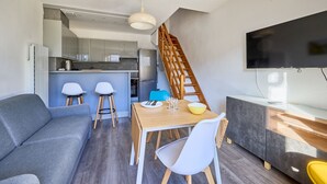 A compact living space with a kitchenette, a dining area, and a TV mounted on the wall. - L'Ancre Bleue - Duplex à 10min de la plage (Courseulles-sur-Mer)