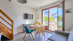 A modern living room with a wooden staircase, a dining area with a table and chairs, a flat-screen TV mounted on the wall, and a sliding glass door leading to an outdoor patio. - L'Ancre Bleue - Duplex à 10min de la plage (Courseulles-sur-Mer)
