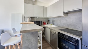 A modern kitchen with a built-in microwave, stove, and dishwasher. There are two white chairs and a small island with a sink. - L'Ancre Bleue - Duplex à 10min de la plage (Courseulles-sur-Mer)