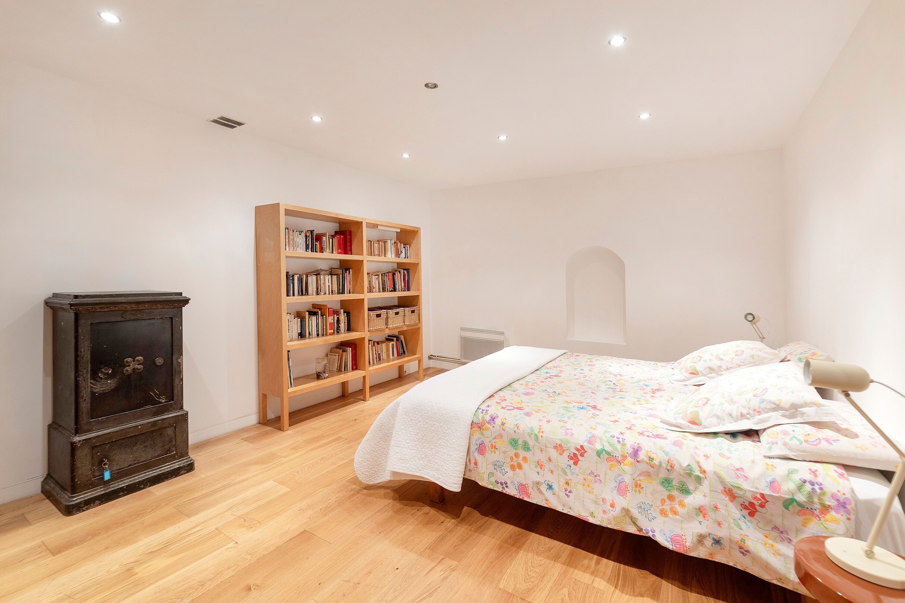 A bedroom with a wooden bookshelf, a bed with a colorful floral bedspread, and a desk lamp.