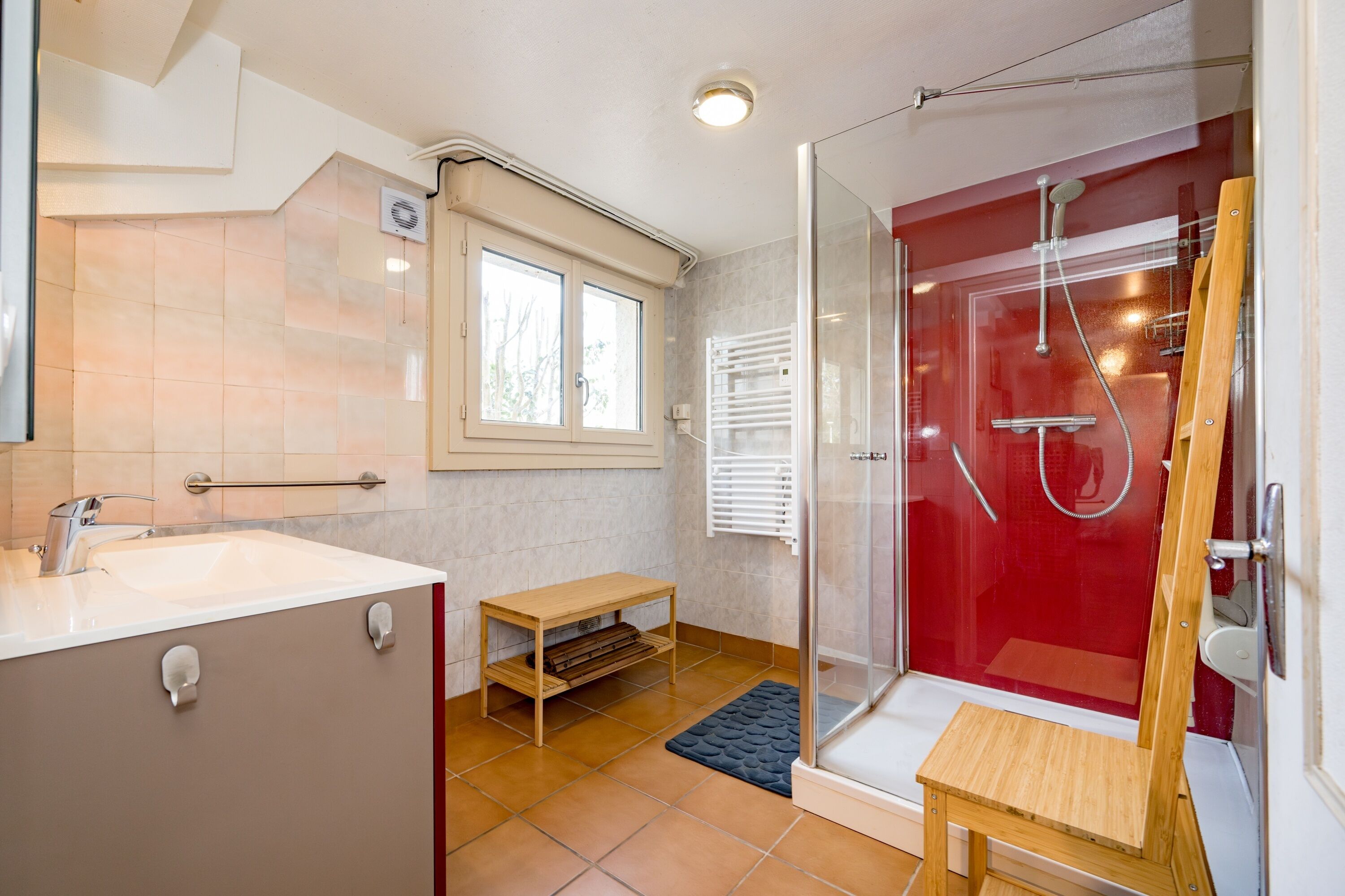 A bathroom with a shower, a sink, a window, and a small wooden table.