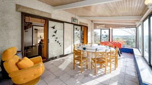 A dining area with a round table and chairs, a yellow armchair, and a view of the outdoors through large windows.