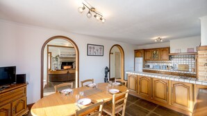 A dining area with a wooden table set for four, surrounded by wooden chairs. There is a kitchen area with wooden cabinets and a tiled backsplash.
