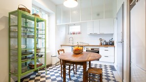 Una cucina con pavimento a scacchi, un tavolo di legno con sedie, una vetrina verde con stoviglie e pensili bianchi.