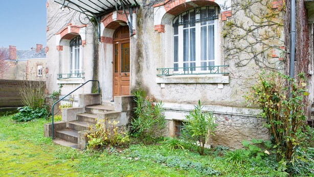 Un edificio storico con facciata in pietra, una porta in legno e un piccolo giardino con dei gradini che conducono all'ingresso.