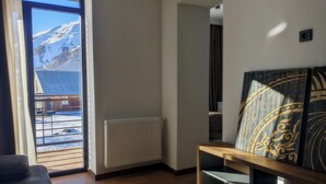 A room with a wooden floor, a radiator, a sofa, a coffee table, and a view of a snowy mountain through a large window. - AGRE Kazbegi (Kazbegi)