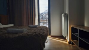 A bedroom with a large bed, a window with a view of snow-capped mountains, a radiator, and a wooden cabinet. - AGRE Kazbegi (Kazbegi)
