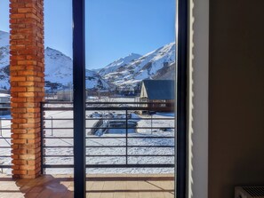 A view through a window with a brick pillar, featuring a snowy mountain landscape, a fenced area, and a building with a sloped roof. - AGRE Kazbegi (Kazbegi)