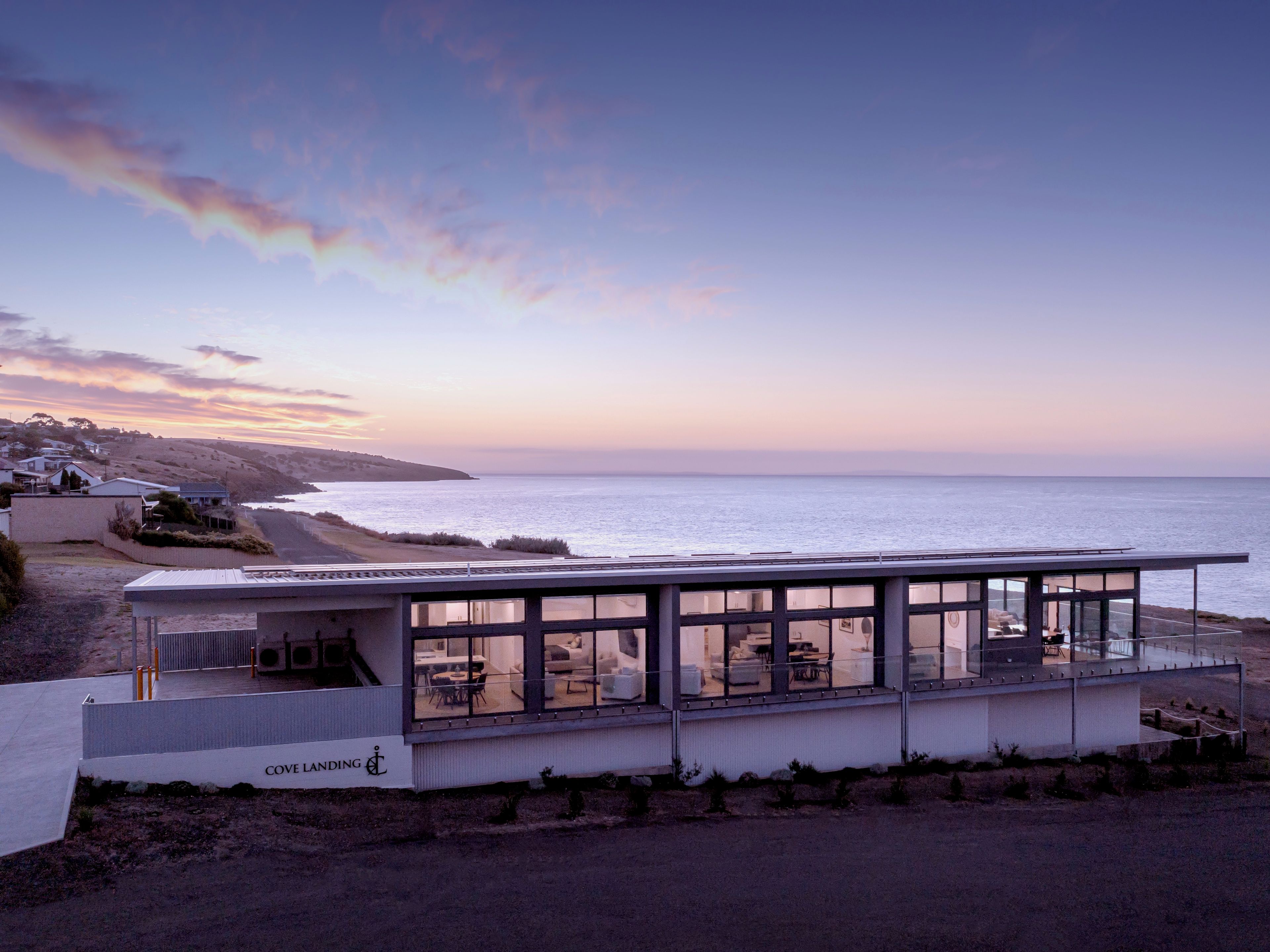 Un bâtiment moderne situé sur la côte, doté de grandes fenêtres offrant une vue sur l’océan, d’un toit plat et d’une enseigne indiquant « COVE LANDING ».