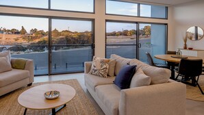 Ein geräumiges Wohnzimmer mit einem großen Fenster und Blick auf eine angelegte Grünfläche. Es gibt ein beiges Sofa, einen runden Couchtisch sowie einen kleinen Esstisch mit Stühlen.